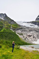 Naklejka na ściany i meble The young girl looks at the Boyabreen glacier, which is the sleeve of large Jostedalsbreen glacier and wild yellow flowers in the foreground. Melting glacier forms the lake with clear water. Norway.