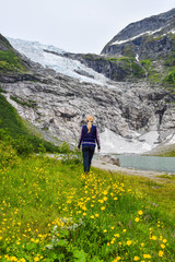 Naklejka na ściany i meble The young girl looks at the Boyabreen glacier, which is the sleeve of large Jostedalsbreen glacier and wild yellow flowers in the foreground. Melting glacier forms the lake with clear water. Norway.