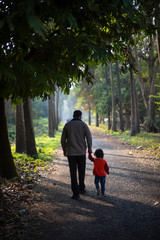 Backside of an Indian brunette father and his baby boy in winter garments walking in a forest path in winter afternoon in natural green background. Indian lifestyle and parenthood.