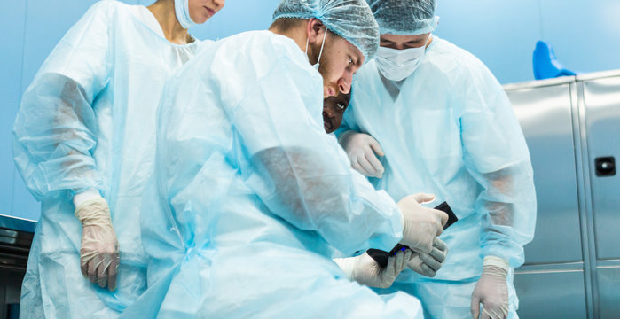 A team of doctors in uniform and medical masks looks at a surgical plan on an electronic tablet