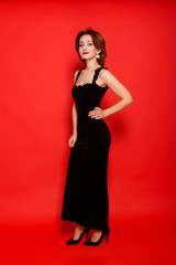 Beautiful, young woman in a black dress and black shoes posing standing on a red background.