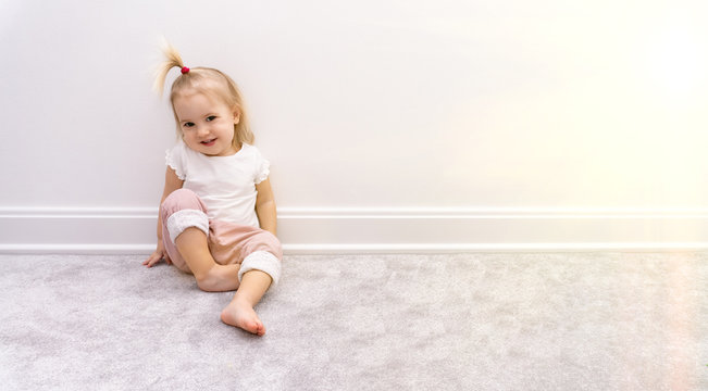 Cute bare foot baby girl in pink trousers sits near white wall on grey rug, smiles and closes eyes from sun, good background, copy space