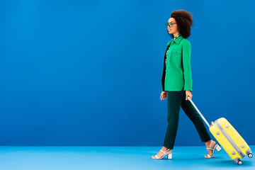 side view of smiling african american woman holding travel bag on blue background