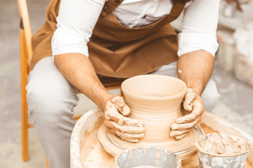 A young male potter is engaged in craft in his workshop on a potter's wheel and makes a clay product