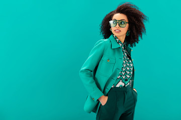 stylish african american woman in jacket with hands in pocket looking away isolated on turquoise