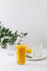 glass of delicious yellow smoothie on white surface near green plant isolated on grey