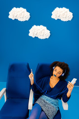 high angle view of african american sitting on seat, listening to music and holding smartphone on blue background with clouds