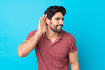Young handsome man with beard over isolated blue background listening to something by putting hand on the ear