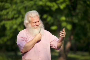 Stylish senior blond man in casual wear looking at smartphone as mirror and scratching his beard by comb and smiling. Closeup portrait of handsome person on sunlit green blur natural background.