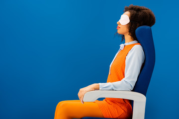 Fototapeta premium side view of african american with sleeping mask sitting on seat and sleeping isolated on blue