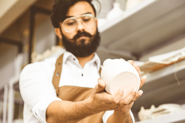 A young attractive male potter with a beard and mustache works in his workshop. Holds and inspects...