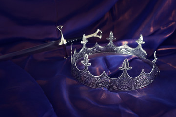low key image of beautiful queen/king crown and sword over dark royal purple delicate silk. fantasy medieval period