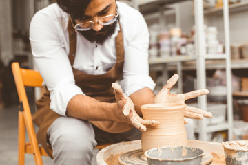 A young male potter is engaged in craft in his workshop on a potter's wheel and makes a clay product