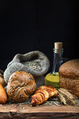A bun, croissant, bread , wheat ear, olive oil and grain in a bag lie on a wooden rustic table against a dark background. The concept of focus. Vertical orientation. Low key. Focus concept