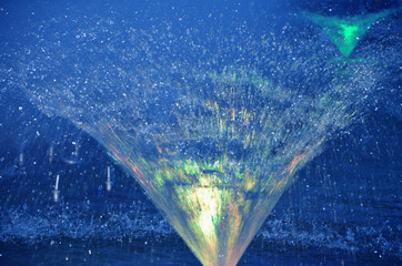 A spray of water from a fountain illuminated in the evening.