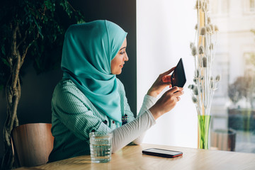 Beautiful young arabic girl in hijab posing for camera at cafe with digital tablet near the window.