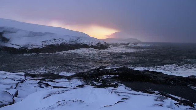 Picturesque sunset on the Barents Sea. Snow flies into the camera. Raging waves hit a rocky snowy shore. The orange sun and blue sky reflect on the surface of the water. Natural background.