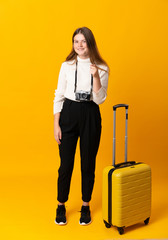 Full body of traveler teenager girl with suitcase over isolated yellow background points finger at you