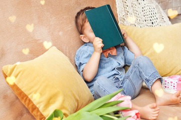A little boy sits at home on his sofa with a book about love dreams and hiding behind a book