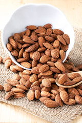 Closeup almonds nut are poured from white bowl on wooden table.