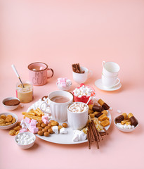 Hot chocolate drink in mugs, sweets assortment of marshmallows, chocolates and cookies on pink background