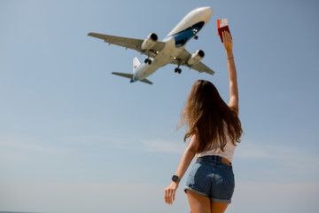 a slender girl in denim shorts and a white t shirt has her arms outstretched passport in hand near the airport and is watching the plane land in anticipation of a trip to Thailand
