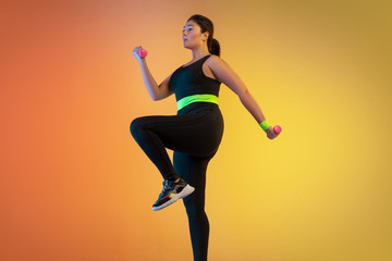 Fototapeta na wymiar Young caucasian plus size female model's training on gradient orange background in neon light. Doing workout exercises with the weights. Concept of sport, healthy lifestyle, body positive, equality.