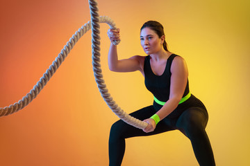 Young caucasian plus size female model's training on gradient orange background in neon light. Training upper body with the fit ropes. Concept of sport, healthy lifestyle, body positive, equality.