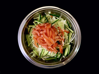 Green salad of cabbage, cucumbers and tomatoes, on a black background. Cutting vegetables in a metal bowl. Finely chopped vegetables for salad. Concept: vegetable diet, healthy nutrition.