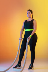 Young caucasian plus size female model's training on gradient orange background in neon light. Posing confident with the fit ropes. Concept of sport, healthy lifestyle, body positive, equality.
