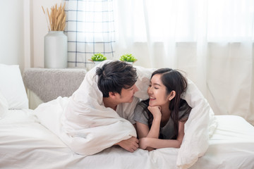 Happy young asian married couple covered with soft warm white blanket together on comfortable bed in bedroom