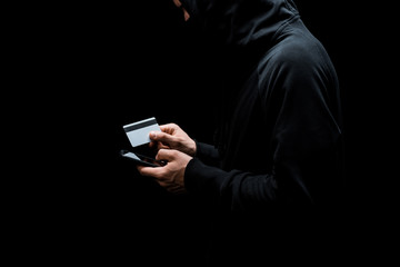 cropped view of hacker in hood and mask using smartphone and holding credit card isolated on black