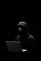 young hacker in hood using laptop isolated on black