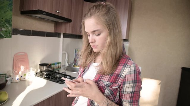 Young married couple in the kitchen. A girl with a tattoo writes a message on her mobile phone to her lover on the background of her husband who washes the dishes.