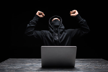 excited hacker in mask celebrating triumph near laptop isolated on black