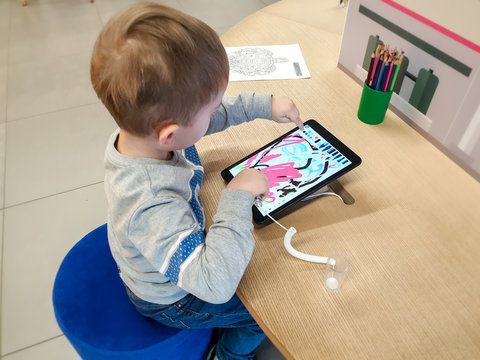 Image of cute 3 years old little toddler boy sitting behind the desk and drawing with stylus on digital tablet computer