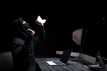 emotional hacker holding tissue near laptop and computer monitors with blank screen isolated on black