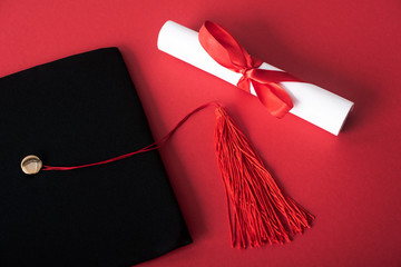Top view of diploma with beautiful bow and black graduation cap with tassel on red background