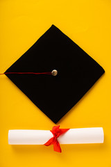 Top view of diploma with beautiful bow and black graduation cap on yellow background