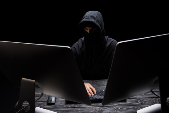 hacker in mask sitting near computer monitors isolated on black