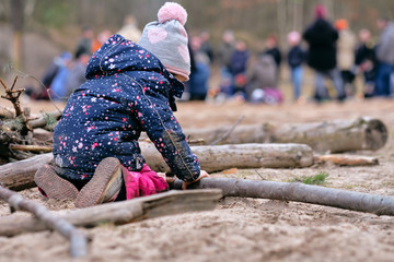 Rear view of a 4 year old child girl in warm clothing kneeing on the ground  and building something with wooden branches in front of a group of people. Seen in Germany in February