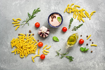 Obraz na płótnie Canvas Raw pasta and ingredients for cooking. Fusilli, tomatoes, basil, olive oil, pink salt, pepper, rosemary and garlic on a gray concrete background. Top view, flat lay