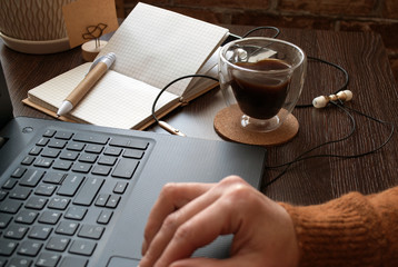 Cropped view of woman sitting with cup of coffee behind wooden table with planners, stationery and laptop .   Glass cup in the shape of a heart and double glass in female hands. 