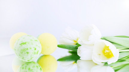 Naklejka na ściany i meble A green and yellow salt bomb on a white gray background is reflected, next to white tulips. Mineral makeup. Banner. Spa care. Banner. Copy space