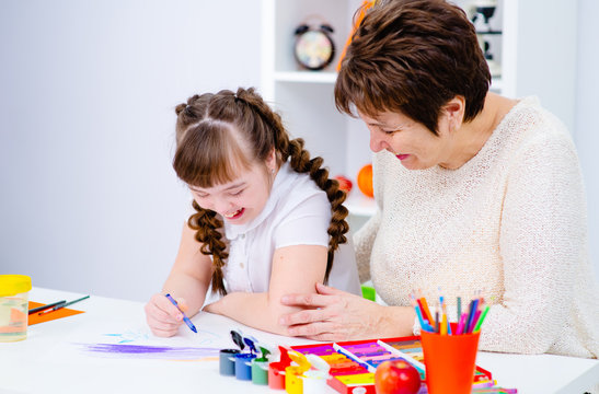 A girl with down syndrome draws at home next to the teacher.