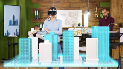 Architect woman wearing VR headset is looking at holographic projections of building a city