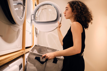 Side view of beautiful girl loading clothes into washing machine in self-service laundry