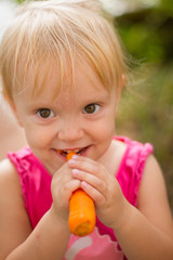 Pretty blond toddler girl eating fresh carrot outdoors on a sunny summer day