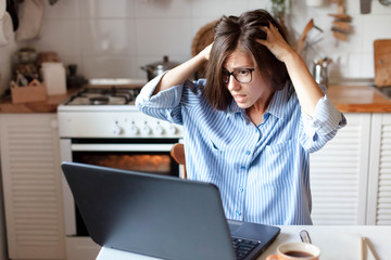 Stressed woman working from home office. Freelancer has trouble with laptop and the Internet. Workplace in cozy kitchen. Concept of female business and career, housekeeping.