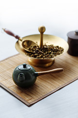Obraz na płótnie Canvas a green Chinese teapot with a wooden handle stands on a white table, behind stands a gold stand with a spatula for tea with green tea
